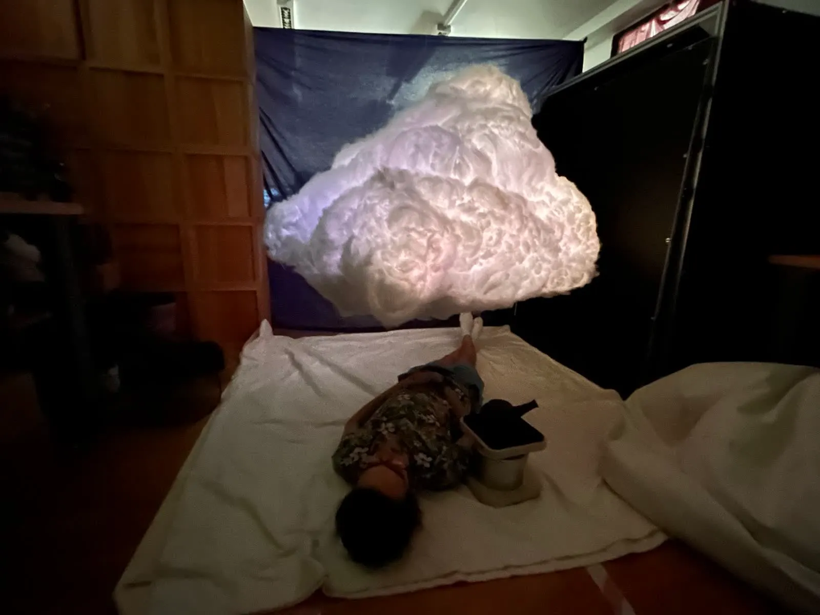 A person lies on white sheets on the floor beneath a large glowing cloud of white fibre, in a dark room with a blue backdrop.