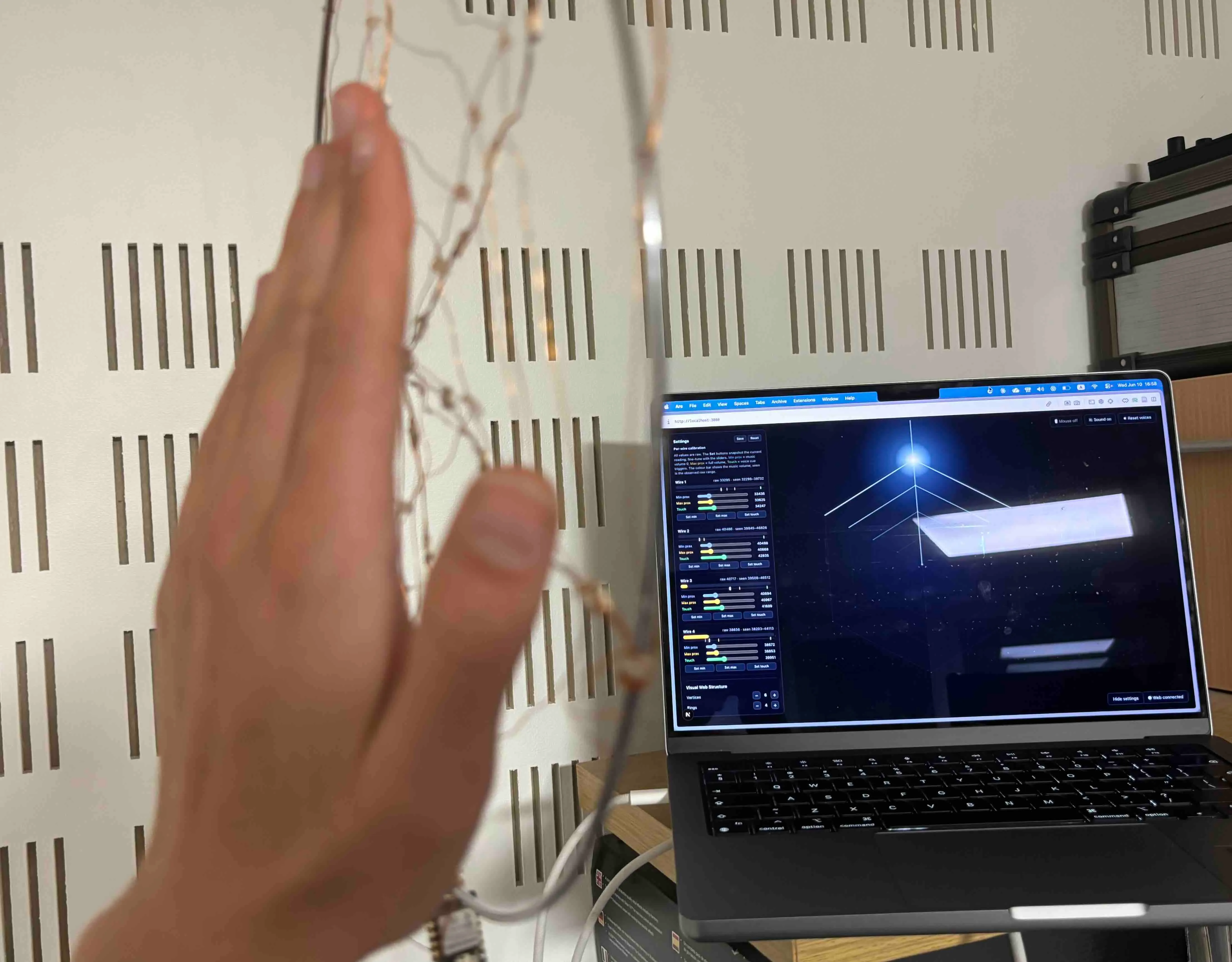 A hand hovering beside the copper-wire web, with the Sensitive Webs interface on a laptop behind it.