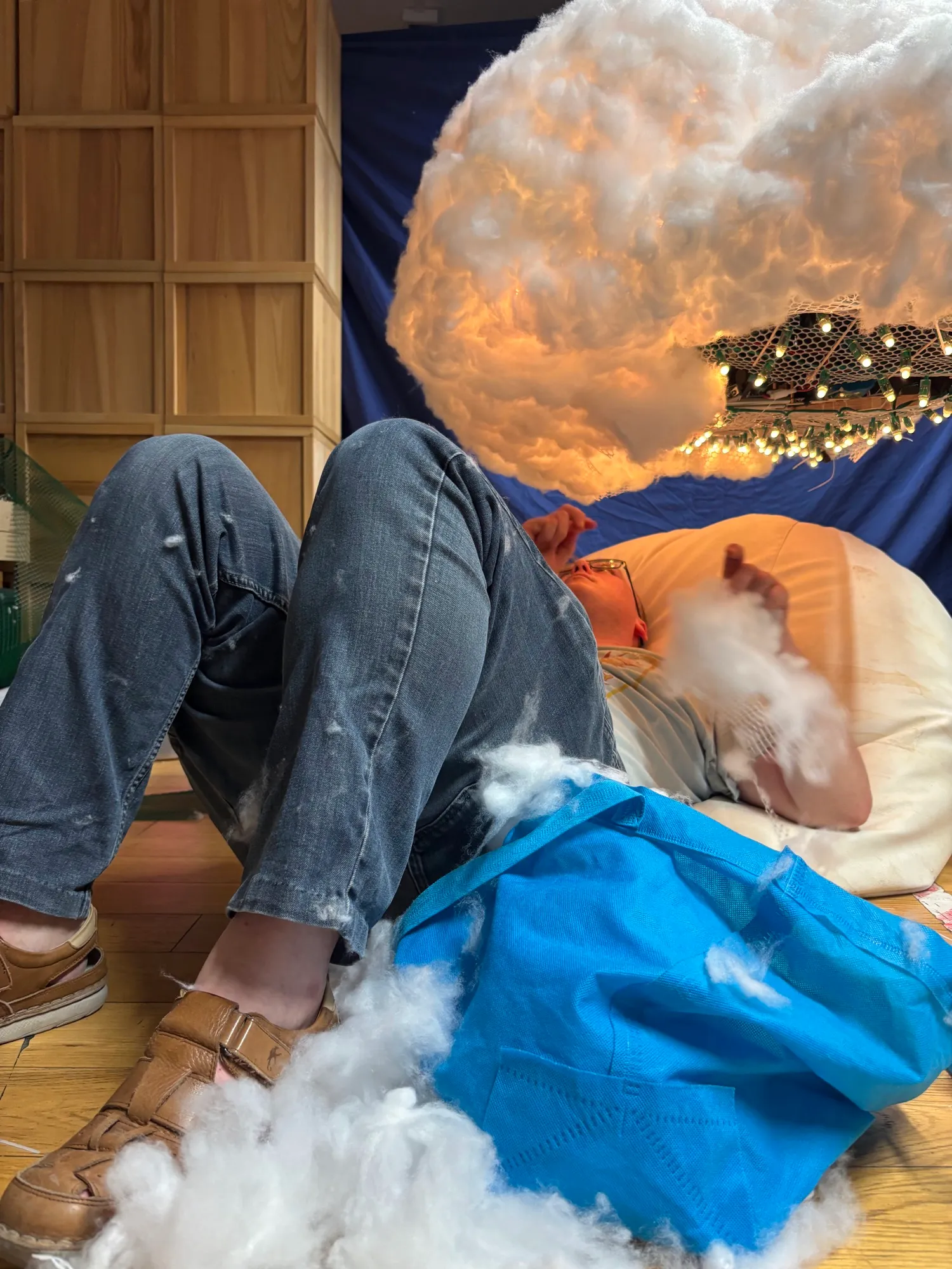 Lying on the floor under the hanging cloud, reaching up to shape it.