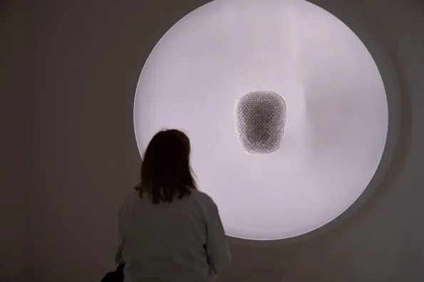 A woman seen from behind, sitting before a large glowing white disc with a small, mesh-covered chest-like object breathing at its centre.