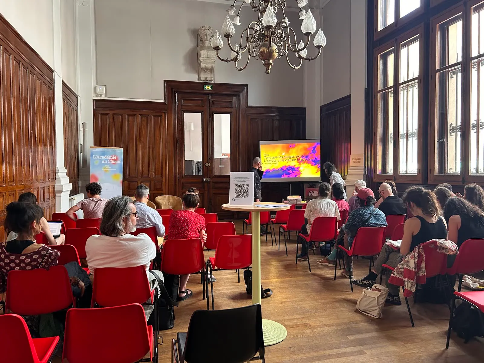 The audience on red chairs in a high-ceilinged room with a chandelier, facing the screen