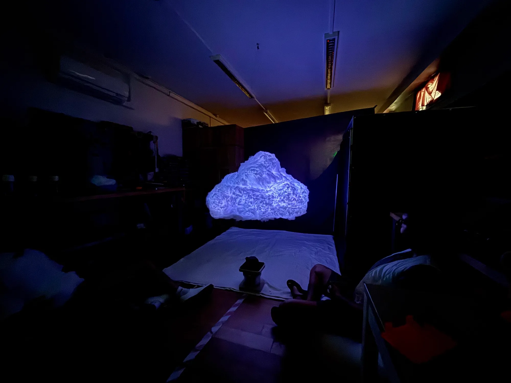 The whole room from above: the cloud glowing over sheets, a small tea table and people sitting on the floor.
