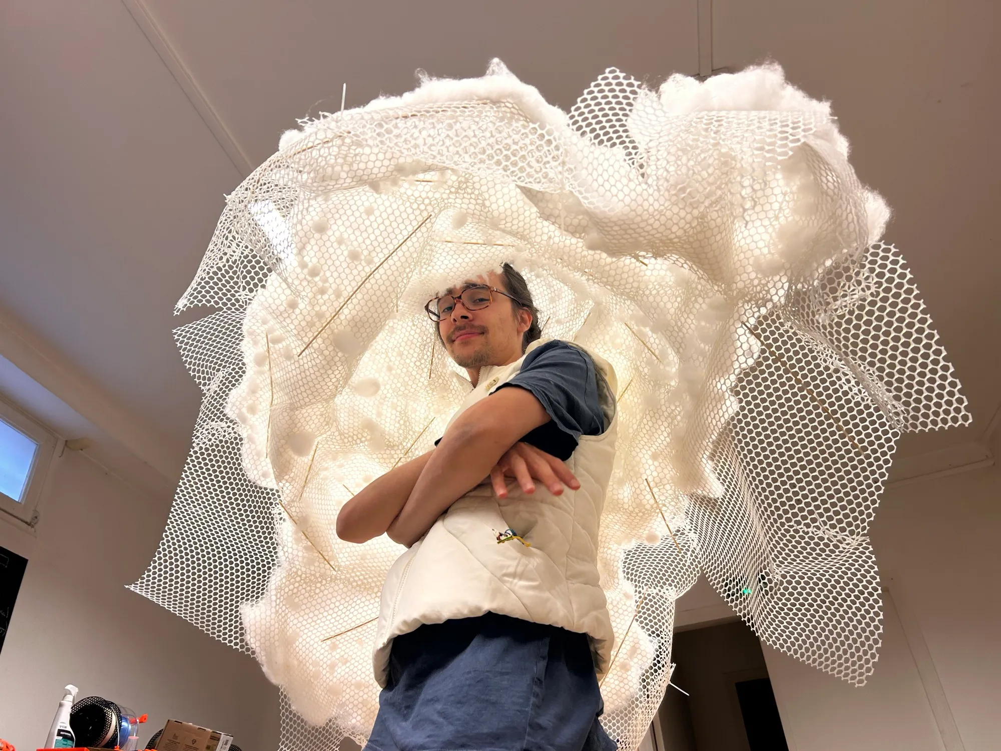 Zéphir stands under the hanging white mesh shell, stuffed with cotton and pinned with skewers, wearing it like a giant cloud hat.